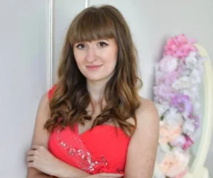 Молода жінка з каштановим волоссям і чубчиком у червоній блискучій сукні схрестила руки